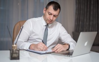 Businessman in suit writing notes at desk with laptop, reflecting modern office life.